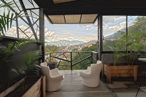 A balcony with two white chairs, potted plants, and a view of a cityscape and mountains. - Luxury Suite | Rooftop + Jacuzzi | Modern Coliving (Medellín)