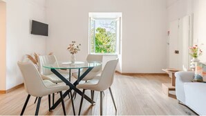 A modern dining area with a glass table and white chairs, a flat-screen TV on the wall, and a window with a view of greenery.