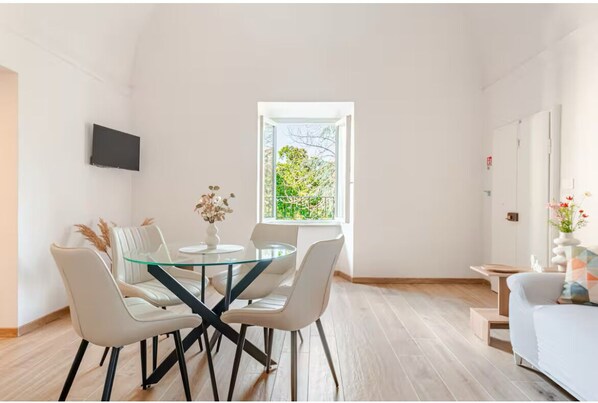 Ein moderner Essbereich mit Glastisch und weißen Stühlen, einem Flachbildfernseher an der Wand und einem Fenster mit Blick auf Grünflächen.