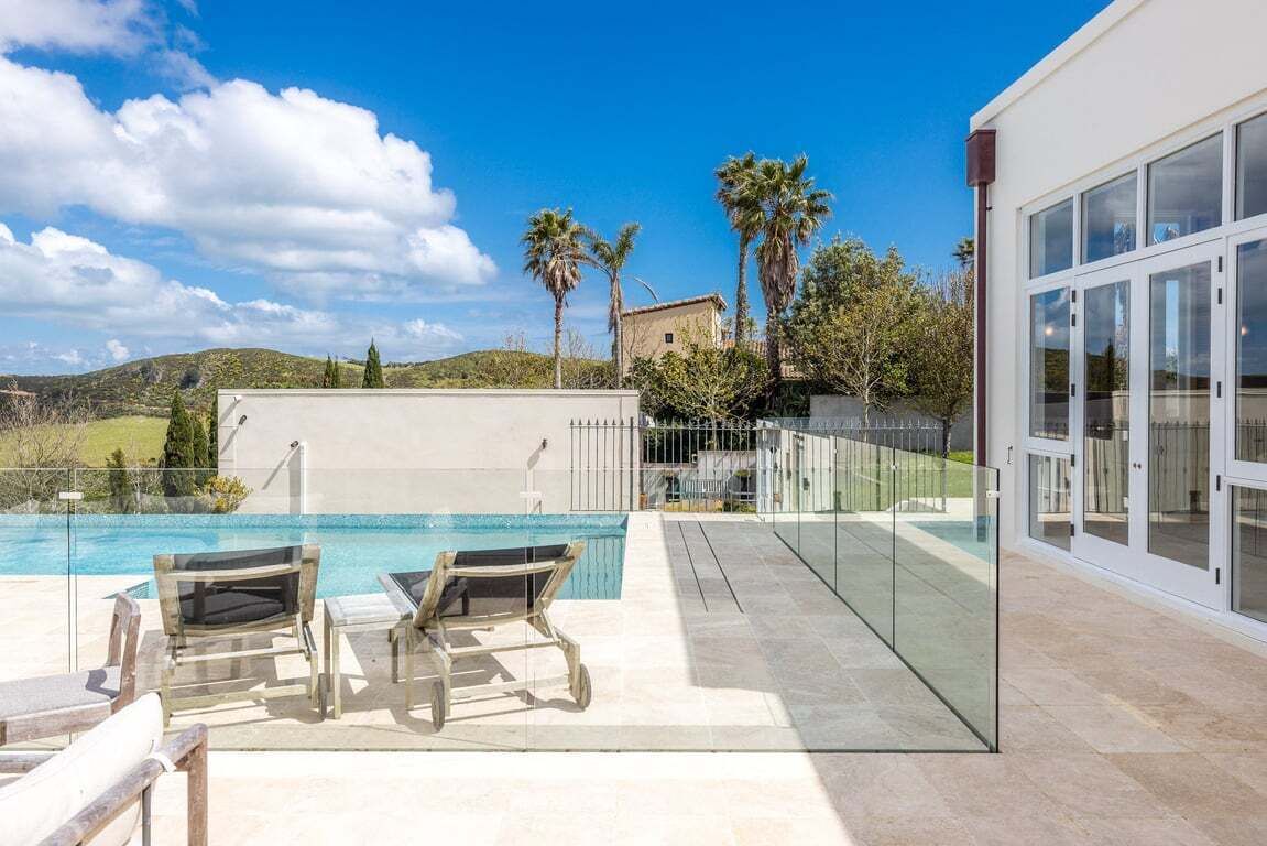 Un área de piscina moderna al aire libre con barandillas de vidrio, dos sillones de descanso y vistas al paisaje circundante, incluyendo colinas y palmeras.