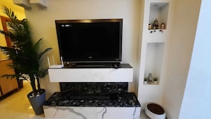 Un téléviseur à écran plat posé sur un meuble blanc avec un socle en marbre, une plante en pot sur la gauche et une étagère avec de petits objets décoratifs sur la droite.