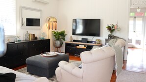 Ett vardagsrum med en platt-TV, en grå soffa, en vit fåtölj, en svart TV-bänk och en krukväxt.