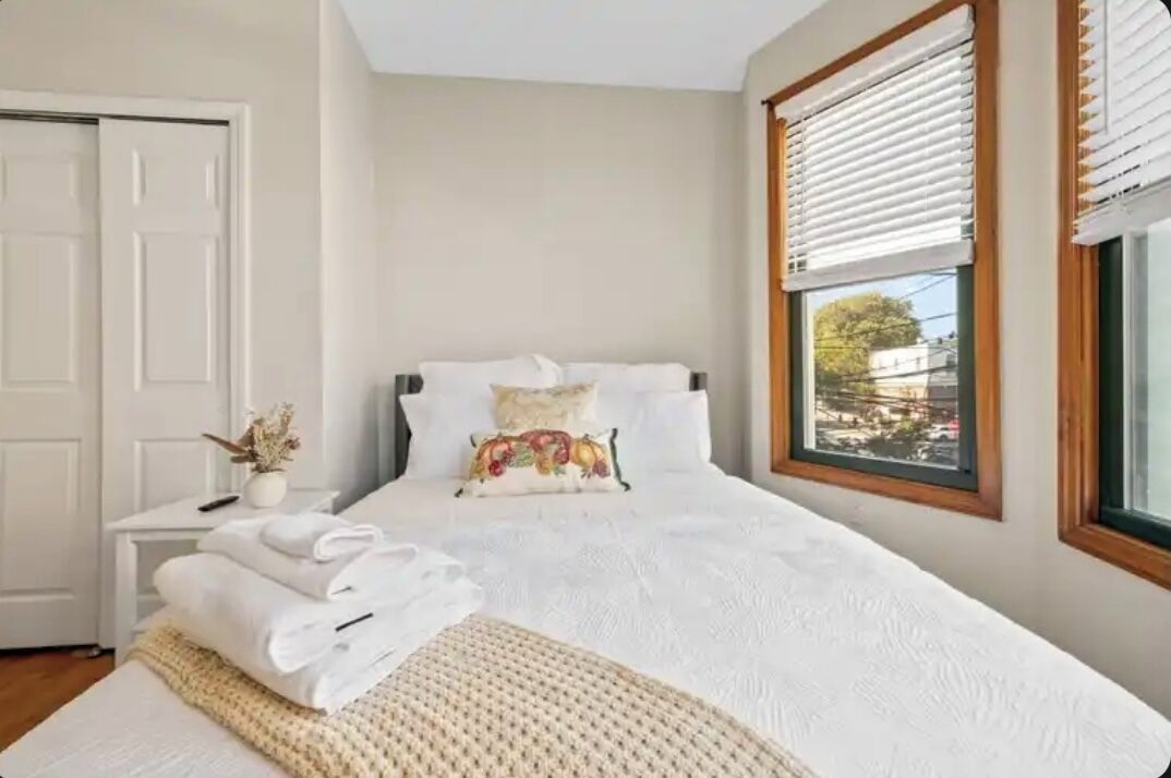 A neatly made bed with white linens, a bedside table with a vase of dried flowers, and a window with blinds.