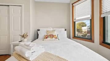 A neatly made bed with white linens, a bedside table with a vase of dried flowers, and a window with blinds.