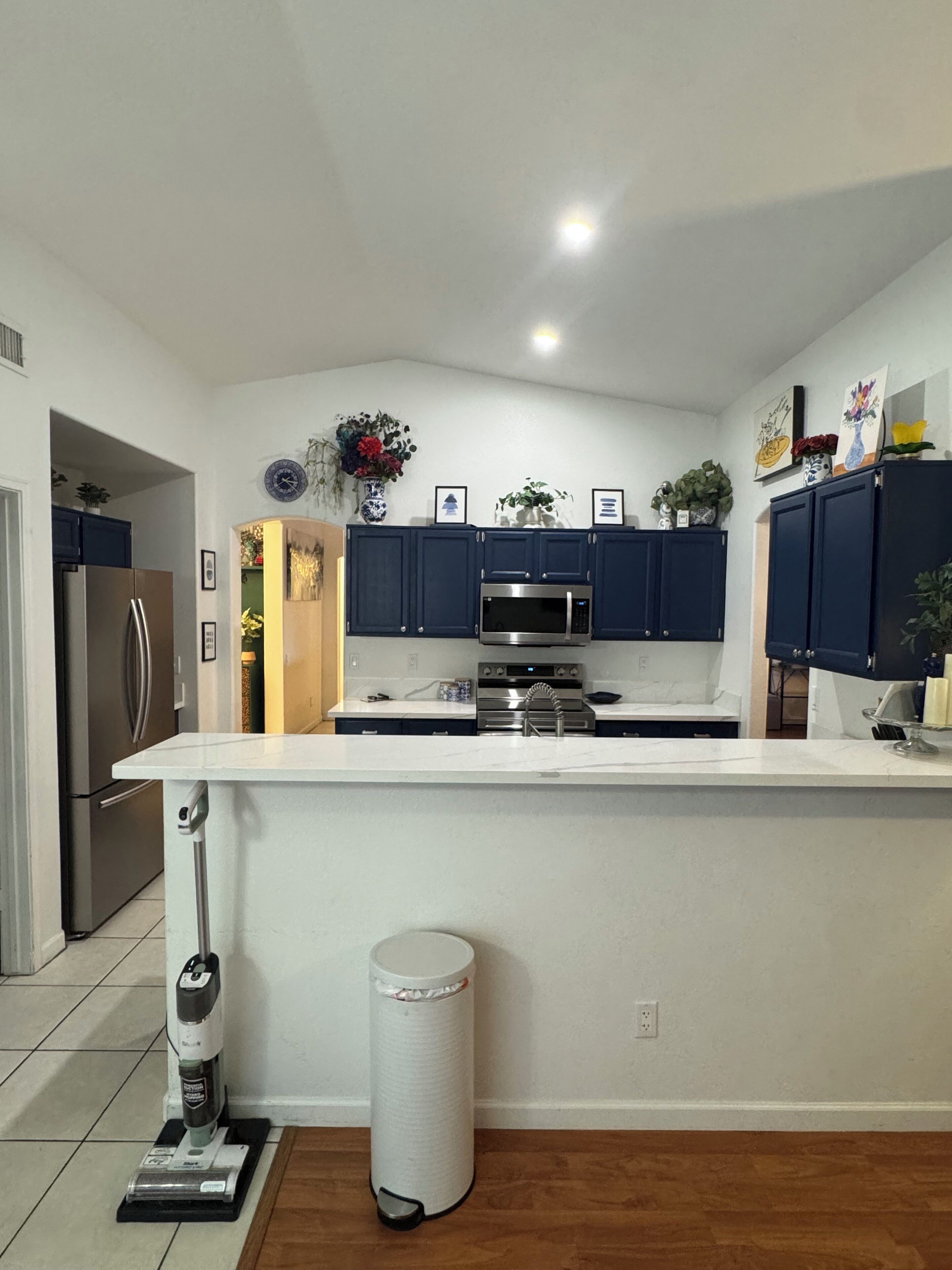 A modern kitchen with dark blue cabinets, a stainless steel refrigerator, a microwave, and a sink. There are decorative items on the countertop and a trash can in the corner.