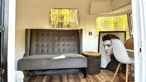 Un dormitorio pequeño con cama de cabecera capitonada, un taburete tejido y un retrato enmarco.