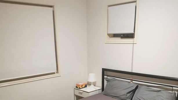 A single bed with a grey mattress and pillows, a bedside table with a lamp, and a window with blinds. - Shared house with other guests near shopping center and theme park (Upper Coomera)