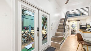 Un interior moderno con una escalera que conduce a un segundo piso, una zona de cocina y un gran ventanal corredizo que da a un área exterior con una estructura verde.