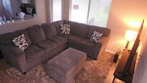 A living room with a sectional sofa, a matching ottoman, and patterned throw pillows. There is a flat-screen TV on a stand to the right, and a window with blinds in the background.