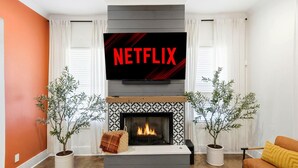 Ett vardagsrum med en öppen spis, en tv som hänger ovanför och visar "NETFLIX", och två krukväxter placerade på varsin sida.