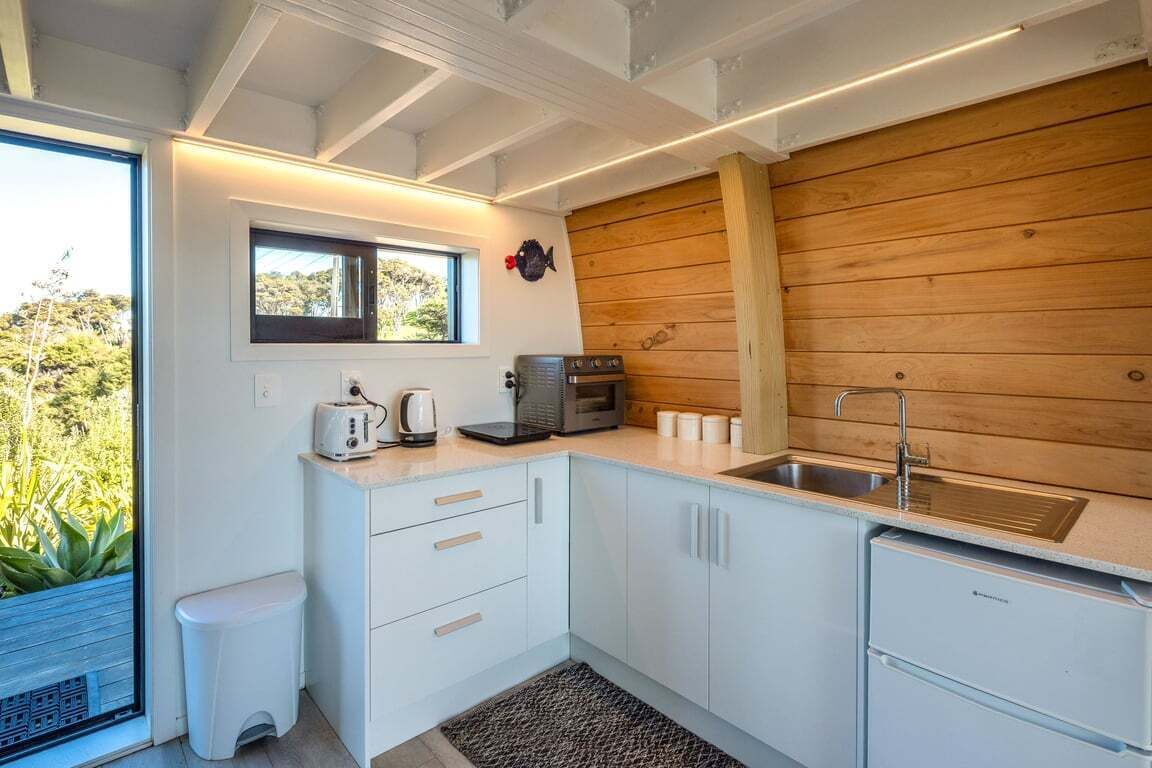 Cocina compacta con armarios blancos, pared de madera, un lavabo y ventana con vistas a la vegetación.