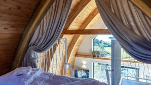 A cozy attic bedroom with wooden beams, a bed, a window with curtains, and a view of the outdoors.