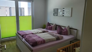A bedroom with a bed, a crib, a window with blinds, and wall art. - Modern, barrier-free apartment with garden access and terrace (Plauen)