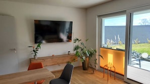 A modern living room with a flat-screen TV mounted on the wall, a wooden bench, and a dining table with chairs. There are potted plants and a small table with a candle. A large window provides a view of the outside. - Modern, barrier-free apartment with garden access and terrace (Plauen)