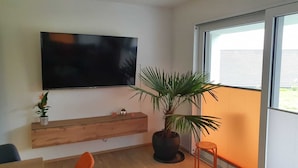 A modern living room with a flat-screen TV mounted on the wall, a wooden console table, a potted plant, and a window with a view of a building. - Modern, barrier-free apartment with garden access and terrace (Plauen)