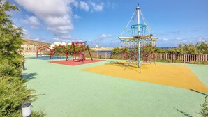 Un terrain de jeux avec des balançoires et une structure d’escalade, entouré de verdure et sous un ciel dégagé.