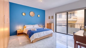 Une chambre moderne avec un grand lit, un mur d’accent bleu, une armoire en bois, une table de chevet, une commode et un balcon avec vue.