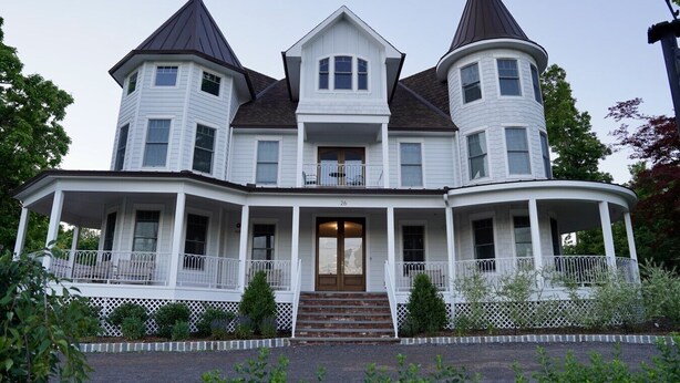 A large, white, two-story house with a wrap-around porch, multiple windows, and two prominent towers.