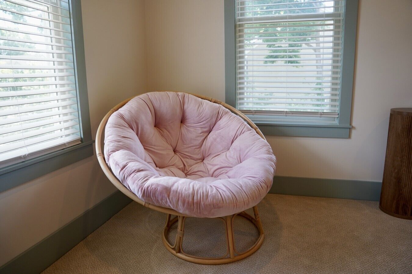 A corner room with a pink chair, two windows with blinds, and a wooden floor.