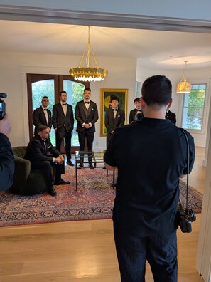 A group of men in suits standing in a room with a large window, a chandelier, and a patterned carpet.