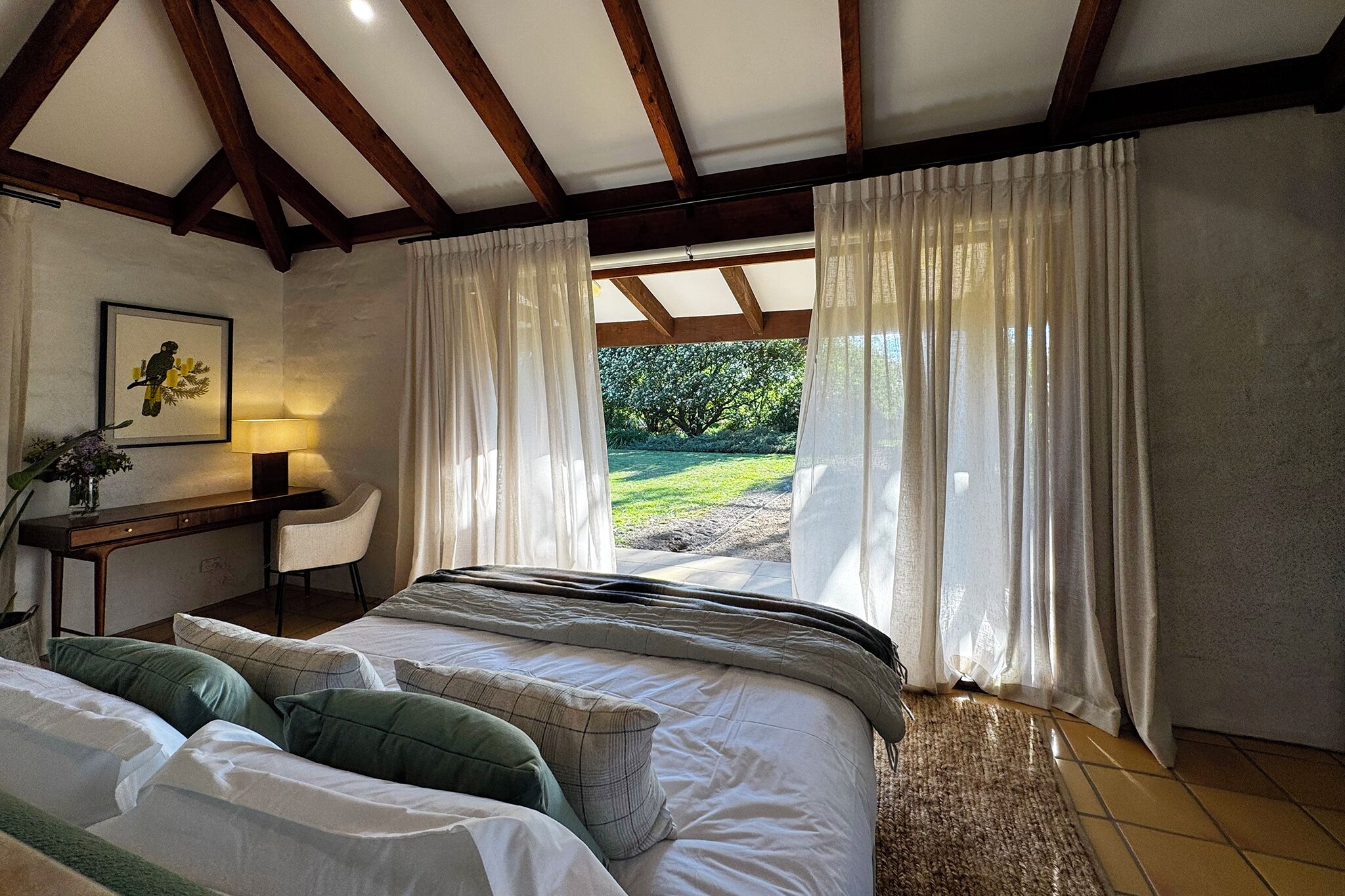 A bedroom with a bed, a desk, and a chair. There is a window with curtains and a view of greenery outside.