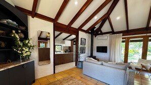 Een ruime woonkamer met een hoge plafond, donkere houten balken, een grijze bank, een salontafel en een keuken die zichtbaar is door een open doorgang.