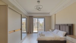 A modern bedroom with a large bed, a nightstand, a wooden dresser, and a view of the city through the balcony doors. - 4-bedroom villa in palm hills compound with garden (Giza Governorate)