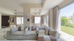 A modern living room with a large sectional sofa, a stone accent wall, and large windows offering a view of a garden. - 4-bedroom villa in palm hills compound with garden (Giza Governorate)