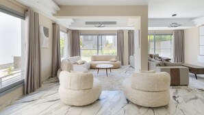A spacious living room with a marble floor, a beige sofa, a round coffee table, and a large window with curtains. - 4-bedroom villa in palm hills compound with garden (Giza Governorate)