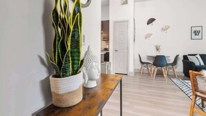 Ein modernes Wohnzimmer mit einem Holztisch, einem Blumentopf und einer Buddha-Statue. Es gibt einen Essbereich mit einem weißen Tisch und Stühlen, sowie ein schwarzes Sofa mit einem gemusterten Überwurf.