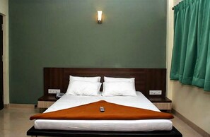 Un lit bien fait, avec du linge de lit blanc et un plaid marron, une table de chevet avec une lampe, et un mur vert agrémenté d’un luminaire.