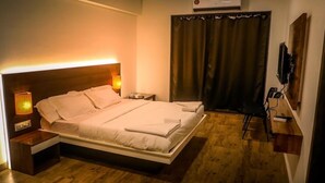 A neatly made bed in a hotel room with a wooden floor, a nightstand with a lamp, a TV mounted on the wall, and a window with curtains. - Hotel Samudra City (Alibag)