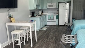 A compact kitchen with white cabinets, a microwave, and a refrigerator. There is a dining area with a table and two stools. The room has a ceiling fan and a bed with a blue bedspread. - BB On The Bayou  (Gautier)