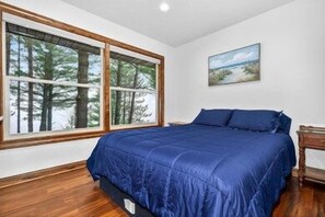 A bedroom with a large window, a bed with blue bedding, and a painting on the wall.