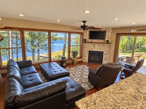 A spacious living room with a large window overlooking a lake, a fireplace, and a ceiling fan. - Lily Bay Lodge (Mercer)
