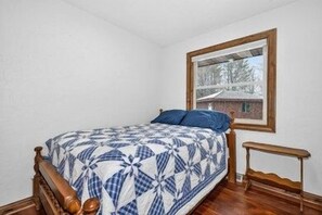 A single bed with a wooden frame and a quilt, a small wooden nightstand, and a window with a view of a house.