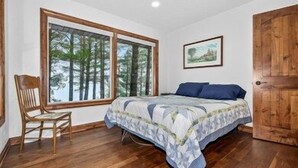 A bedroom with a large window, a bed with a patterned bedspread, a wooden door, and a chair.