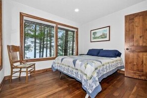 A bedroom with a large window, a bed with a patterned bedspread, a wooden door, and a chair.