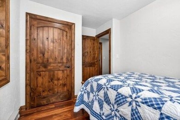 A bedroom with a wooden door, a bed with a blue and white quilt, and white walls. - Lily Bay Lodge (Mercer)