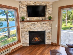 A living room with a stone fireplace, a flat-screen TV mounted above it, and large windows offering a view of a lake and trees.