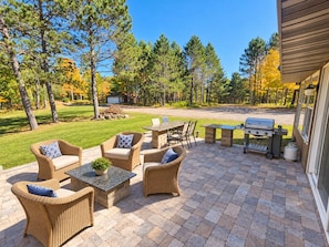 An outdoor patio with wicker furniture, a grill, and a view of a forest. - Lily Bay Lodge (Mercer)