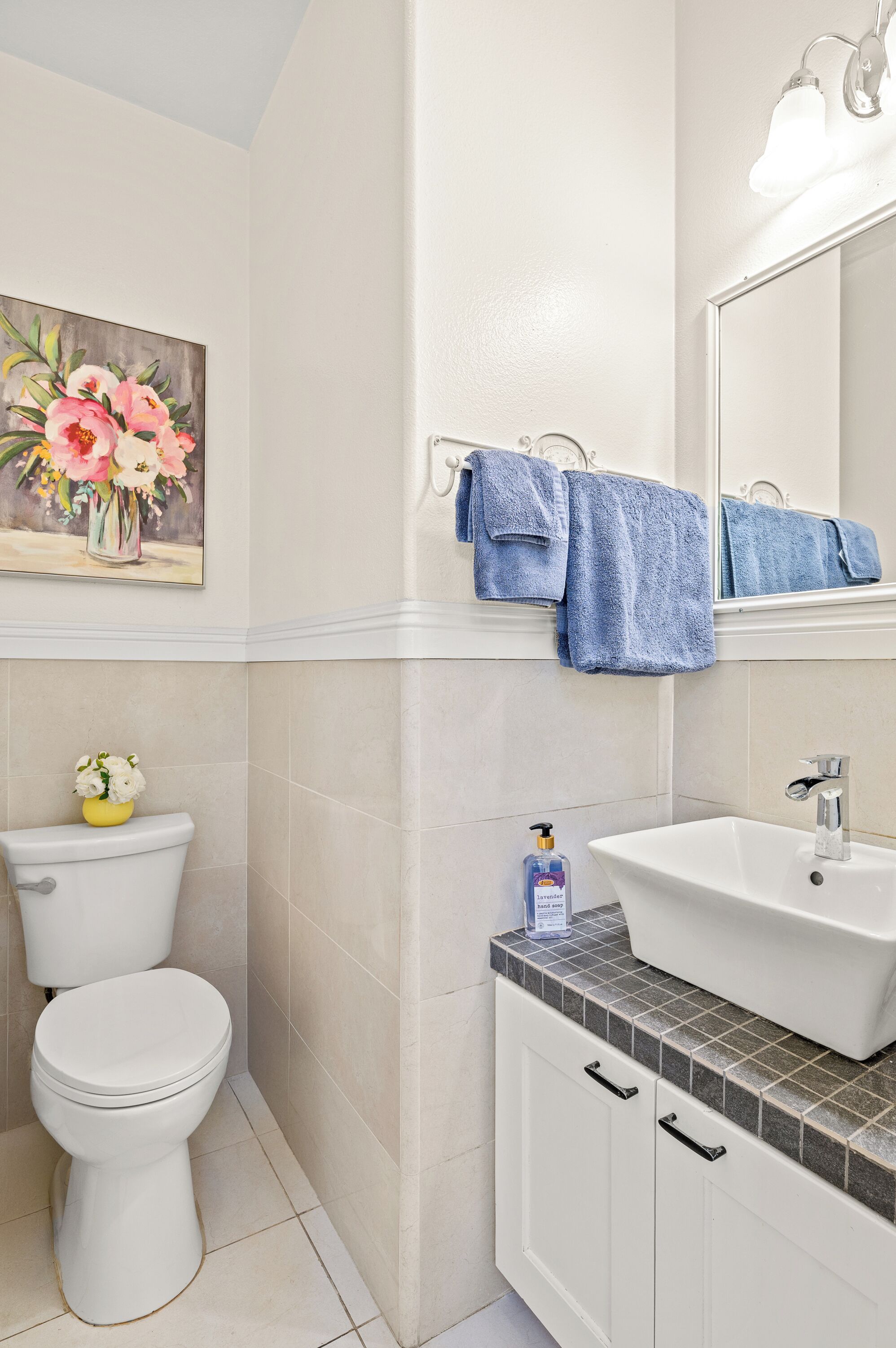 A bathroom with a toilet, sink, mirror, and a painting of flowers on the wall.