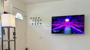 Een woonkamer met een glazen salontafel, een bank, een kamerkleed met dessin, een televisie aan de muur, en een brandblusser.