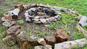 Een achtertuin met een vuurkuil, omringd door houtblokken en rotsen, een houten terras en een picknicktafel.