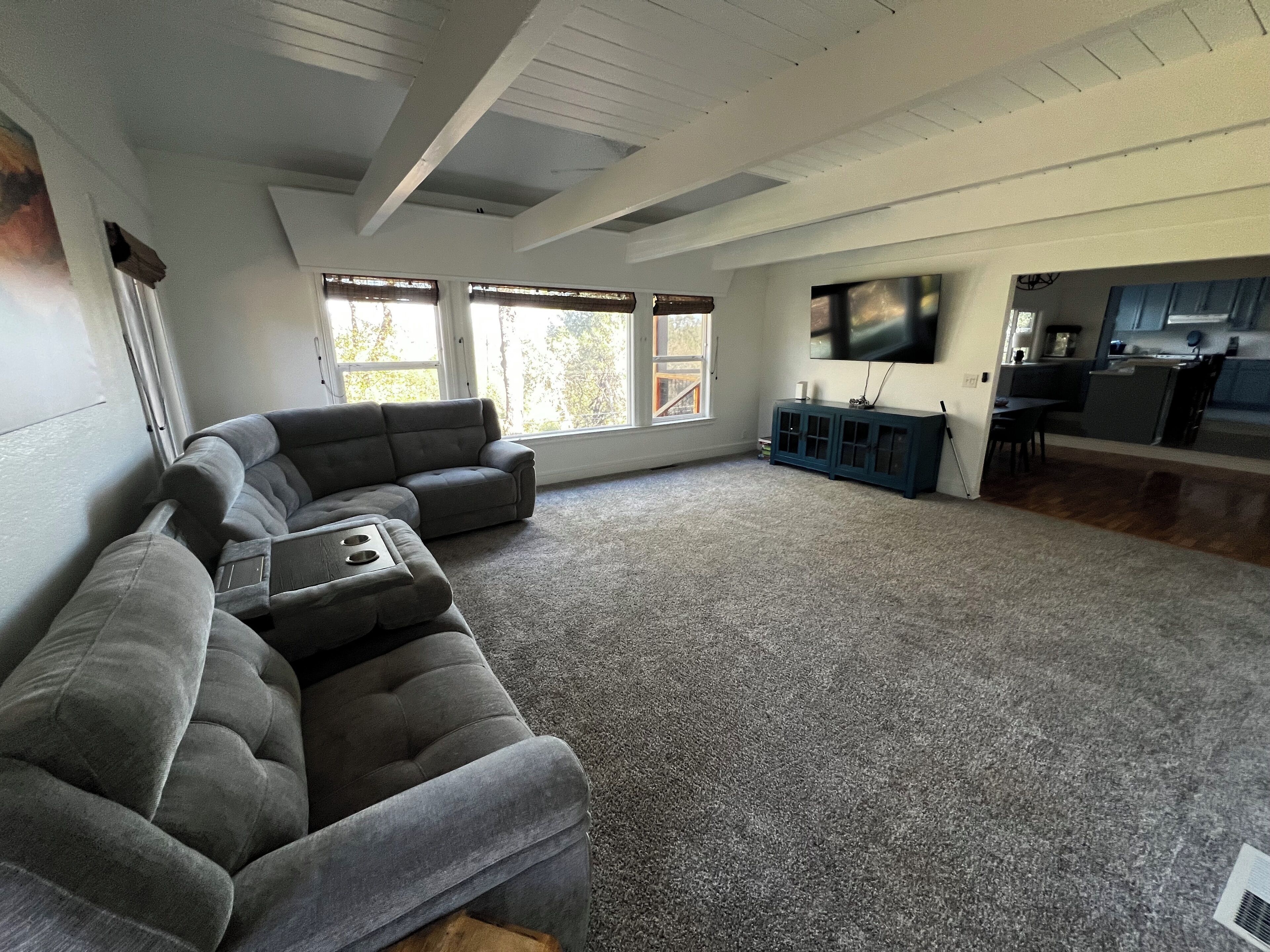 A spacious living room with a grey sectional sofa, a small coffee table, and a large TV mounted on the wall. There are windows with blinds, a kitchen area with cabinets, and a wooden floor.
