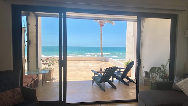 Una stanza con una porta scorrevole in vetro che dà su un balcone con vista sull'oceano e una palma. Sul balcone ci sono due sedie e un tavolino.