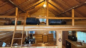 Interior rústico de una cabaña con cama en altillo, una zona de cocina con electrodomésticos básicos y un comedor con dos sillas.