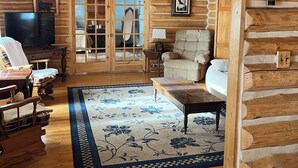 Interior de una cabaña de madera con suelo de madera, alfombra floral, televisor, sofá y sillón.