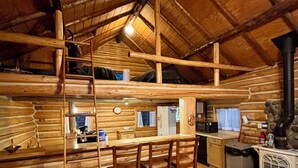 Interior rústico de una cabaña con zona de comedor de madera, un altillo y un estufa de leña.