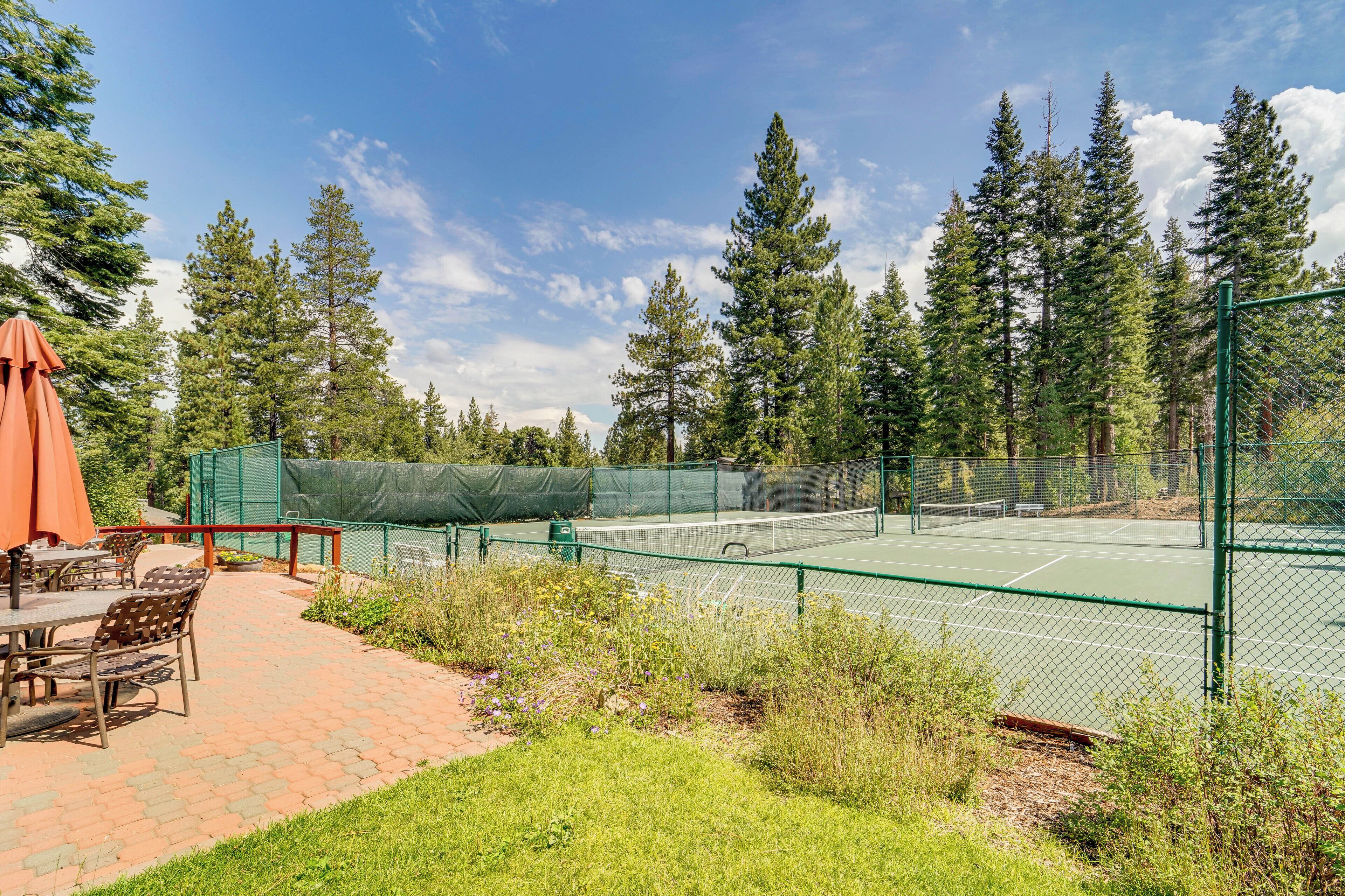 Tenniskenttä, jonka ympärillä on vihreä aita, keskellä on verkko ja sen yhteydessä on patio, jossa on tuoleja ja aurinkovarjo.